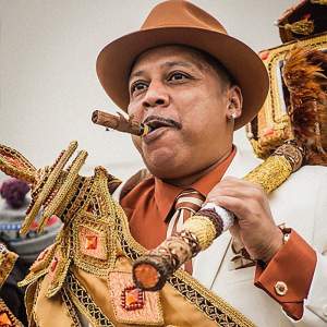 Big Chief Tyrone "Pie" Stevenson wears an ornately beaded sash over a white suit, wearing a brown fedora with a cigar in his mouth. He is a Black man with medium skin tone and a pencil mustache.