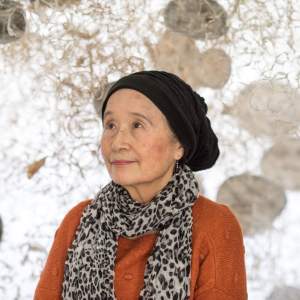Yuriko Yamaguchi stands amidst a sculptural installation, wearing a slouchy black hat, a leopard print scarf and orange sweater. She is a Japanese-American woman with light skin tone.