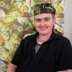 Vee Adams sits in a wooden chair with their hands resting in their lap, and smiles warmly towards us. Vee is white with short black hair dyed yellow at the top and front, and wears a black short-sleeved button up over a fishnet material, with green and white checkered paints. Behind are tiled prints showing magnified fungus structures in green.