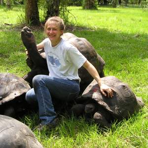 Rachel Berwick sits with her arms around two boulder-sized tortoises. Rachel is white and her dark blond hair is pulled back. She smiles widely and wears a white t-shirt and jeans.