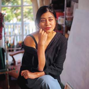 Farah Billah is a Bangladeshi woman with black hair. She wears a black shirt falling off one shoulder and blue pants. She sits with her legs crossed in a studio space. One forearm and elbow rest on the bent knee, and she looks off into the distance with that hand brushing her cheek.