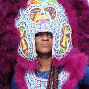 Big Queen Kelly Pearson Boles is a Black Native American Creet woman. She smiles out from under a headpiece that almost completely encircles her face. It is completely beaded with purple, pale blue, white, brown, and pale yellow rhinestones and beads. Fuschia-pink feathers line the head piece and the chest piece on her torso.