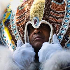 Big Chief Zee Bo Boles looks out from under a beaded head piece, which he holds with both white-gloved hands. He has dark skin and a hint of a five o’clock shadow.