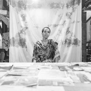 Black and white photo of paris cian, who sits on the far side of a table strewn with images. paris looks steadily at us with a hint of a smile. She has short-cropped hair and wears a dyed wrap. A cloth hanging from above screens much of the studio space behind paris.