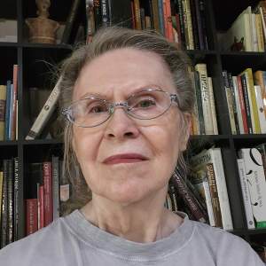 Celia Eberle is a white woman with her dark gray hair pulled back. She wears oval-framed glasses and a gray t-shirt. She looks at us from in front of shelves packed with books and two figurines.