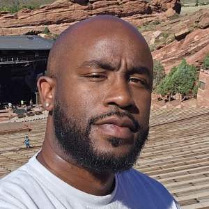 Brandon Felix is a bald Black man with a trimmed beard, and he wears a gray t-shirt. The corners of his mouth lift and he squints slightly as he tips his head to our right. Rows of seats zoom appear to descend steeply to an open-air theater, which is nestled in jutting boulders and scrubby growth on rolling mountains in the distance.