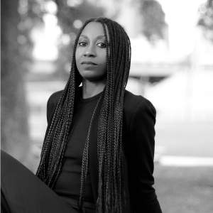 Black and white photograph of Shana M. griffin shows the artist sitting outside and leaning slightly back on her hands. Shana has long braids and wears dark clothing.