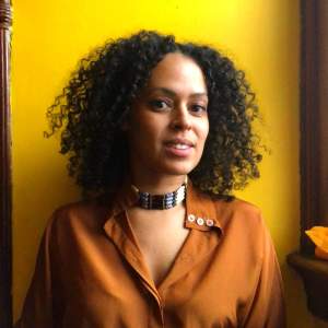 Ana María Agüero Johannes is a Black Hispanic artist with shoulder-length, curly black hair. They wear a wide, beaded choker and an umber-brown blouse, and they look out and smile at us against a canary-yellow background.
