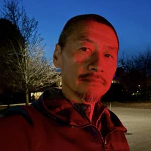Fred H. C. Liang is an artist of Asian descent. He has short-cropped dark hair and a goatee. He is lit with red light, and he cuts his eyes to our left, perhaps taking a selfie with his right hand. Trees are silhouetted against a twilight blue sky in the distance.