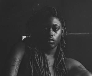 Felicita Felli Maynard looks steadily out at us in this shadowy black and white photograph. Felli has dark-toned skin and long, thin dreadlocks. Parts of dark tattoos are visible on the chest and shoulders.