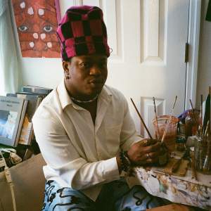Hakeem Olayinka looks at us with a hint of a smile in a studio corner. He sits at a table with painting supplies, with 3 paintbrushes clasped in his hands. He is a Black man with black hair tucked into a tall pink and black checkered hat. He wears a white buttoned shirt with light blue satin pants with swirling black lines.