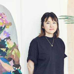 Naomi Kawanishi Reis is an artist of Asian descent. She has brown, shoulder-length hair and bangs, and she wears a navy-blue, short-sleeved shirt and gold necklace with a pendant.