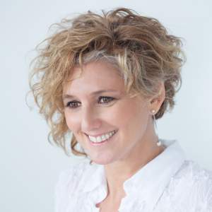 Analia Segal is a Latina artist with short, curly blond hair. She wears a white shirt and smiles as she looks off to our left in front of a white background.