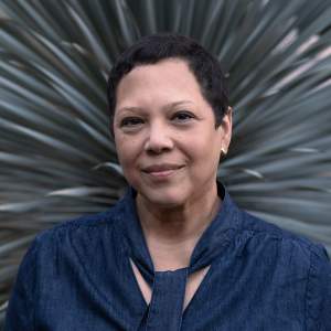 Edra Soto is a Puerto Rican artist with short dark hair. In this headshot, wears a denim shirt with a tied sash on the collar and small gold earrings. She looks and smiles out at us with a blurred palm plant in the background radiating out from her head.
