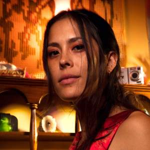 Jade Thiraswas is a Thai-American artist with long black hair, and she wears a vivid red sleeveless garment. She looks slightly down at us from in front of a piece of dimly lit furniture and wall covering.
