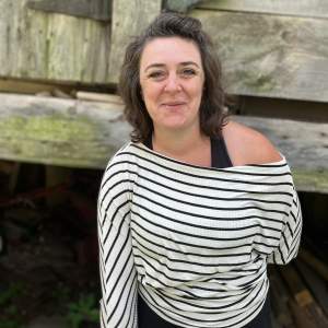 Caitlin Ezell Waugh stands smiling in front of wood paneling, perhaps of an outdoor building. She is a white woman with dark brown hair. She wears a white and black horizontally striped shirt that falls off one shoulder, and she holds one arm behind her back.