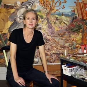 Margaret Curtis sits in a studio with her hands on her knees in front of a detailed painting and a work cart covered with painting supplies. She is a white woman with light skin tone, long grey-blonde hair pulled back, and wears dark jeans and a black tshirt.