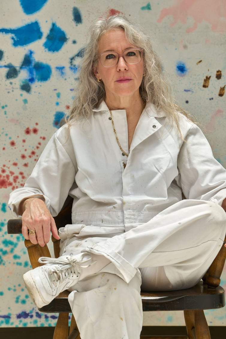 Rebecca Morris sits on a wooden chair wearing a white jumpsuit and sneakers. She is a white woman with light skin tone and long gray hair, with a colorful abstract painting behind her.