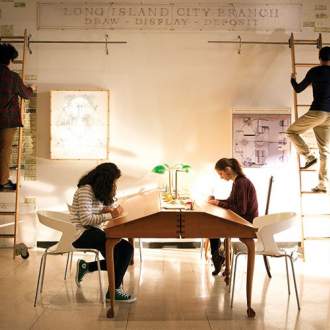 People interacting with art exhibit: two figures on ladders against wall with text on it, and two figures seated at library table