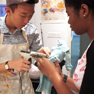 A person in an apron uses a glue gun on a piece of fabric held in the hands of another person.