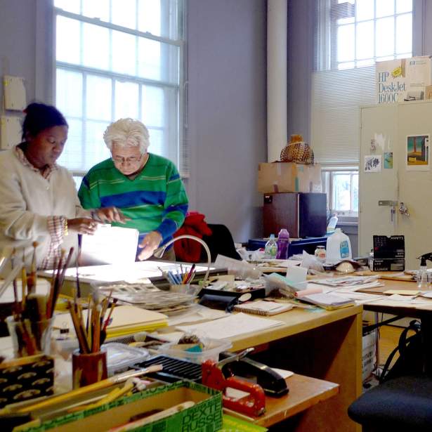 Alex Unthank and Marcos Dimas looking at slides on a lightbox in a room filled with papers, art supplies and storage.