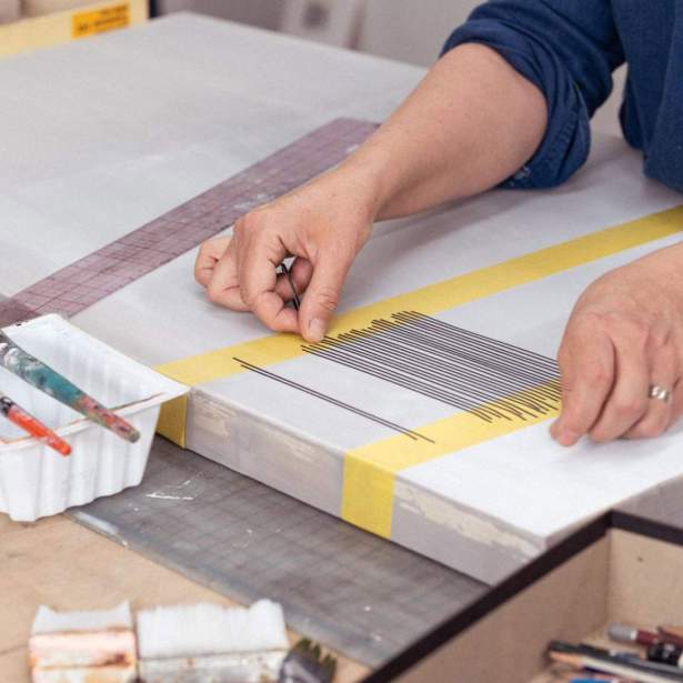 Emily Gherard's hands lay thin black horizontal strips bordered by strips of yellow tape on white silk stretched over a frame. Brushes, pencils and other art supplied are to the side on the wooden table. Gherard is a white woman wearing a denim button-up shirt, and photo is cropped at her chest.