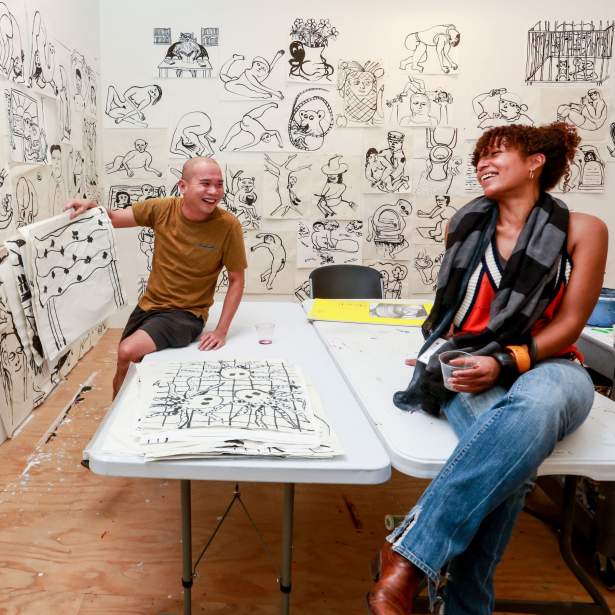Artist Ralph Pugay smiling and showing an ink drawing to laughing artist Cecelia Fernandes, seated on tables, surrounded by black and white ink drawings on the white studio walls