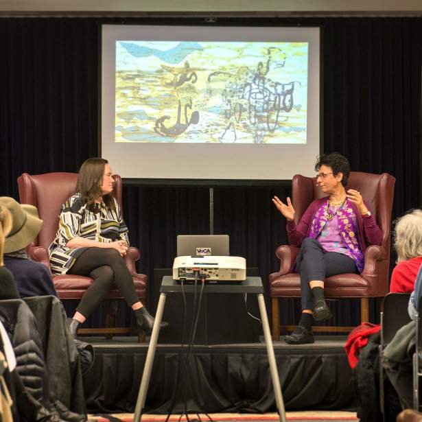 Artist Tara Sabharwal talking to Robin Clark in front of an audience, behind them a screen shows Sabharwal's artwork